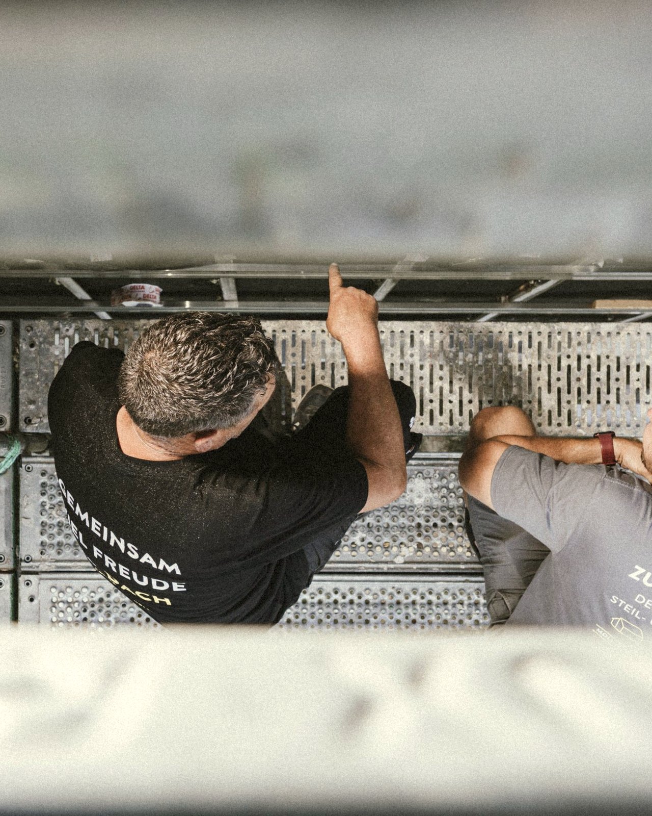 Zwei M-Team-Mitarbeiter aus der Vogelperspektive auf dem Gerüst — Moll-Workwear 'Gemeinsam auf Freude Flach' und 'Zuhausebauer'
